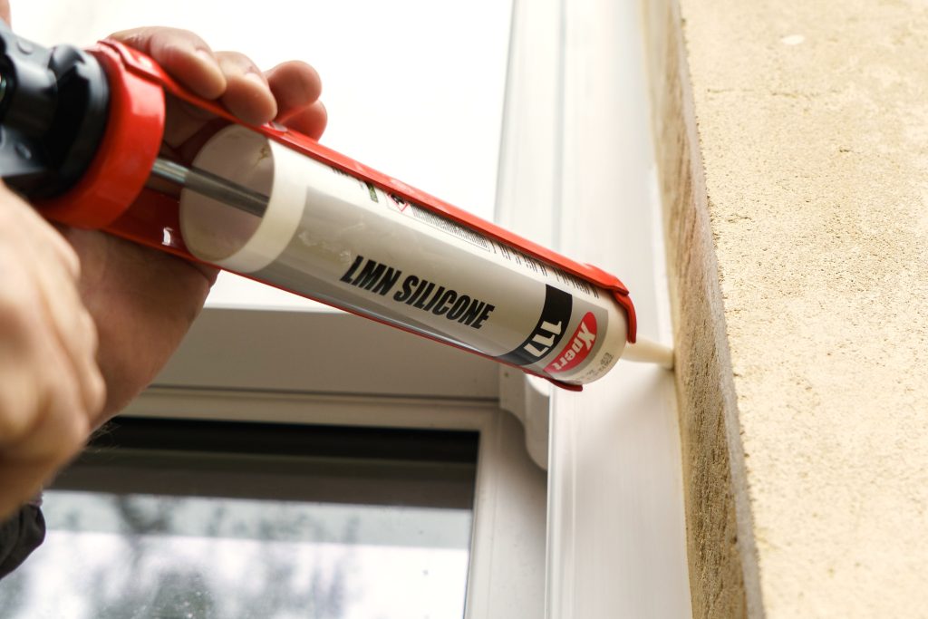 Close-up of a worker applying Xpert 117 LMN Silicone sealant to a white window frame with a red caulking gun.